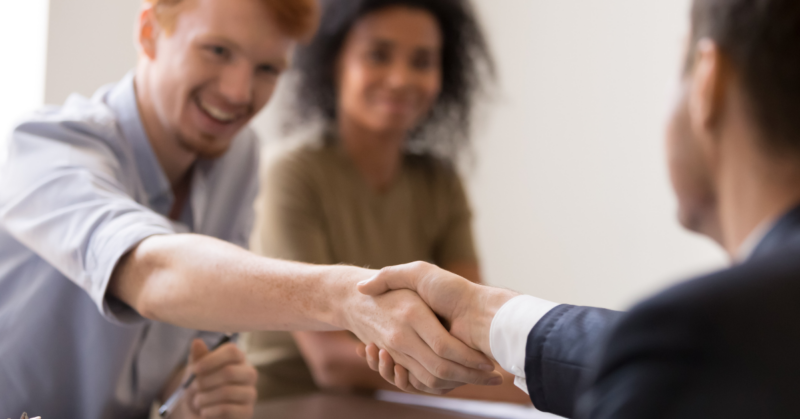 Two people shaking hands across a table in front of a woman.